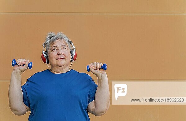 Weißhaarige fettleibige ältere Frau  die mit Hanteln und Kopfhörern trainiert und Musik zur Gewichtsabnahme hört  vororangefarbenem Hintergrund und Kopierraum
