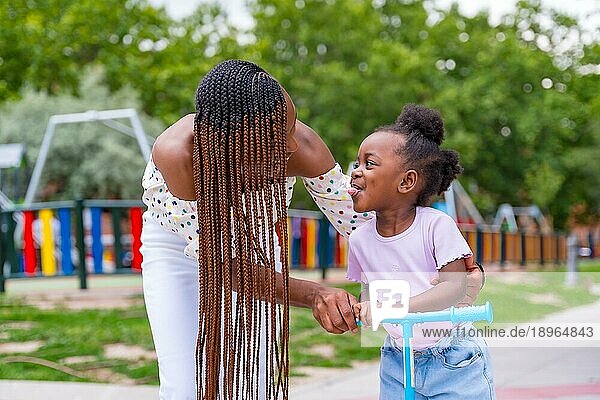 Schwarzafrikanische Mutter hat Spaß mit ihrer Tochter auf dem Spielplatz  die mit dem Skateboard fährt und ihre Zunge herausstreckt