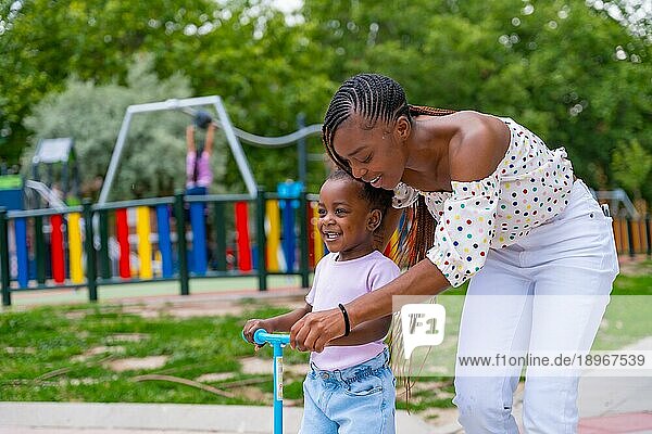 Schwarzafrikanische Mutter hat Spaß mit ihrer Tochter auf dem Spielplatz und lernt  Skateboard zu fahren