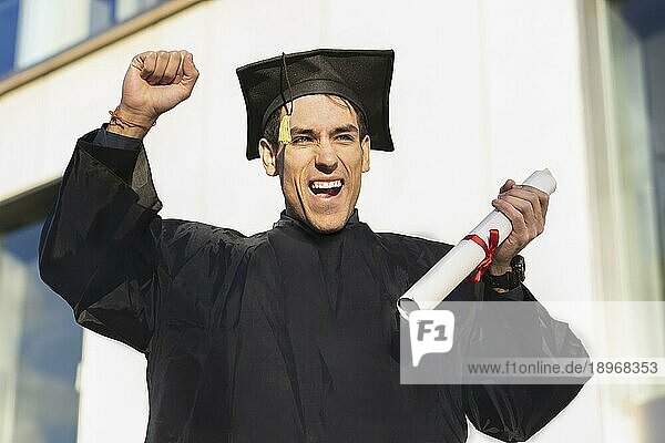 Glücklicher junger Mann in Abschlussrobe  der sein Diplom hält und den Arm hebt
