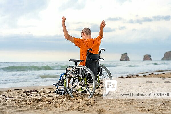 Eine behinderte Person auf dem Rücken in einem Rollstuhl am Strand mit offenen Armen  die die Freiheit des Meeres und der Natur genießen