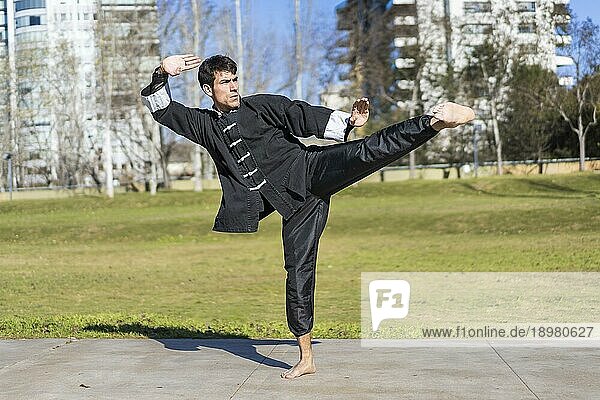 Junger athletischer Kampfsportler  der in einer Kung Fu Uniform Kicks in einem öffentlichen Park übt