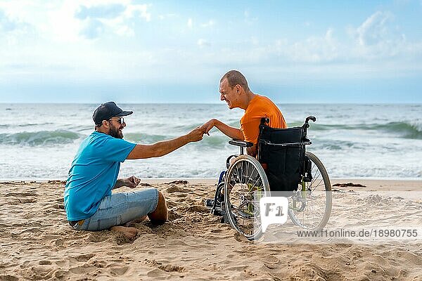 Eine behinderte Person im Rollstuhl am Strand mit einem Freund  der auf das Meer schaut und die Hände hält