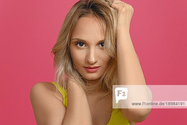 Attraktive junge blonde Frau mit den Händen in den Haaren  die mit einem nachdenklichen  ruhigen Lächeln in Nahaufnahme vor einem roten Studiohintergrund in die Kamera schaut
