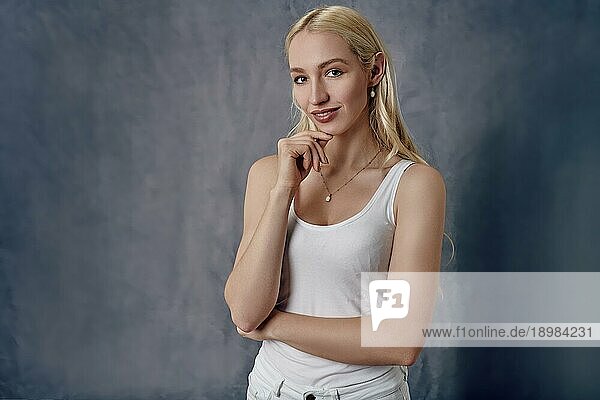 Attraktive  sexy  schlanke  blonde Frau mit langen Haaren  die in einem weißen Sommertop und Jeans posiert und mit der Hand am Kinn in die Kamera lächelt  vor einem grauen Studiohintergrund in einem Dreiviertelporträt
