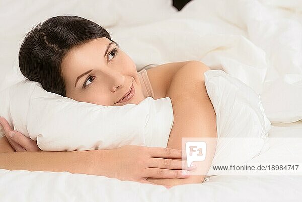 Lächelnde  gesunde  verjüngte junge Frau  die sich im Bett zwischen ihre Kissen kuschelt und der Kamera ein breites  freudiges Lächeln schenkt