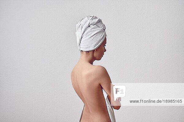 Schlanke junge Frau nach dem Duschen von hinten mit weißem Handtuch über dem Kopf und mit nacktem Rücken. Porträt in halber Länge vor einfachem hellgrauem Hintergrund