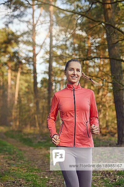 Gesunde  fitte  attraktive junge Frau  die im Wald auf einem Waldweg joggt und sich der Kamera mit einem glücklichen Lächeln nähert  in einem aktiven Lebensstil  Gesundheits und Fitnesskonzept