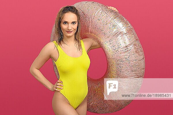 Sexy schlanke junge Frau im gelben Badeanzug  die einen großen aufblasbaren Schlauch über dem Arm hält  während sie mit der Hand auf der Hüfte in die Kamera lächelt  vor einem roten Hintergrund