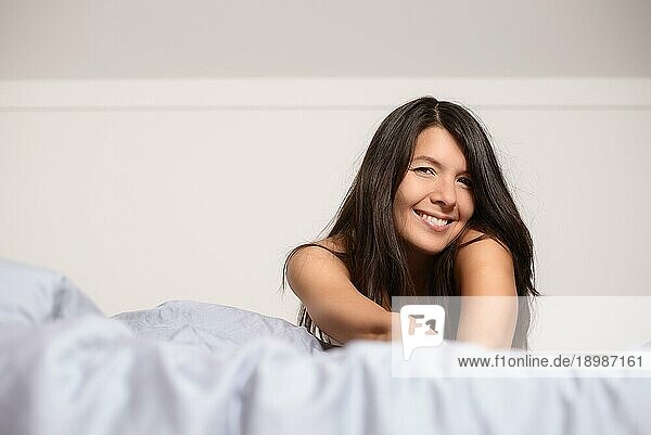 Lächelnde attraktive junge Frau  die einen faulen Tag genießt  auf dem Bauch liegend  sich in eine warme Bettdecke kuschelnd und mit einem glücklichen Lächeln in die Kamera blickend