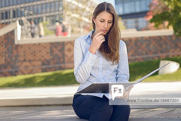 Attraktive Frau  die in einer Mittagspause oder auf einer Reise in eine fremde Stadt in einem Park auf einer Bank im Schatten eines Baumes eine Geschäftsakte liest oder ihre Notizen studiert