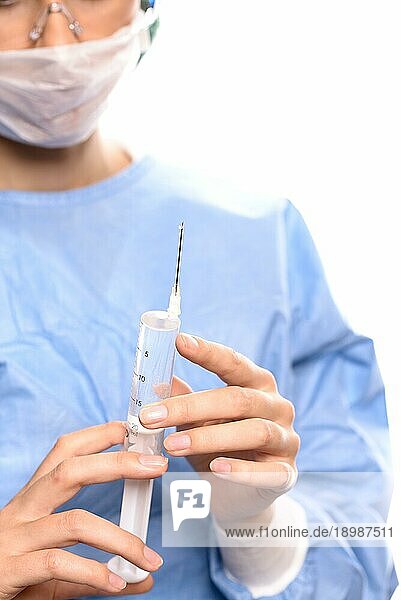 Nahaufnahme der Hände einer Ärztin  die einen blaün Arztkittel  eine Schutzmaske und eine Schutzbrille trägt  während sie die Luft aus einer mit einem flüssigen Medikament gefüllten Spritze entfernt  um eine Embolie zu verhindern