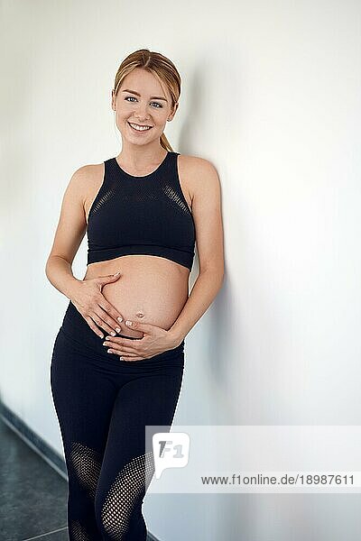 Attraktive  schwangere  freundliche junge Frau  die an eine weiße Wand gelehnt steht und ihre Hände auf ihren nackten Bauch legt und ihren Babybauch streichelt  während sie sich mit ihrem ungeborenen Kind verbindet und glücklich in die Kamera lächelt