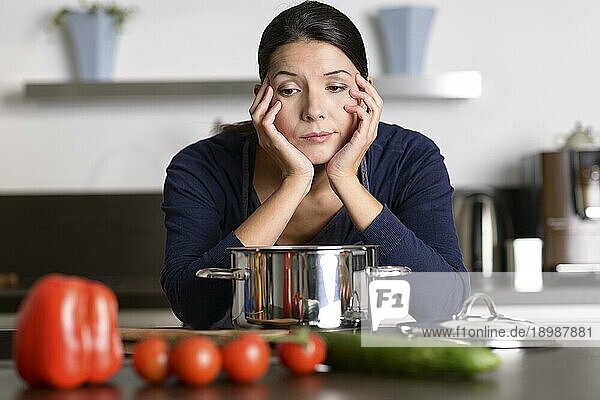 Unmotivierte attraktive junge Frau  die das Abendessen zubereitet und sich dabei auf das Kochfeld stützt und das frische Gemüse mit lustlosem  mürrischem Blick betrachtet  während sie in ihrer Küche in einer Schürze steht