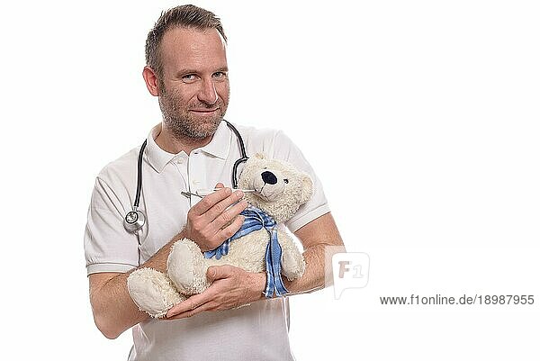 Lächelnde  unrasierte  fürsorgliche Kinderkrankenschwester oder Kinderarzt mit einem Teddybären im Arm  der seine Temperatur mit einem Thermometer mißt  um einen kleinen Patienten während einer Konsultation zu beruhigen
