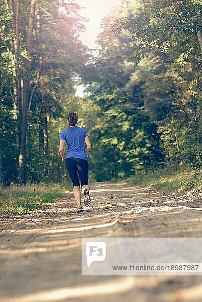Sportliche junge Frau in Sportkleidung  die auf einem Waldweg im Rahmen eines Fitness und Wellnesskonzepts von der Kamera weg joggt