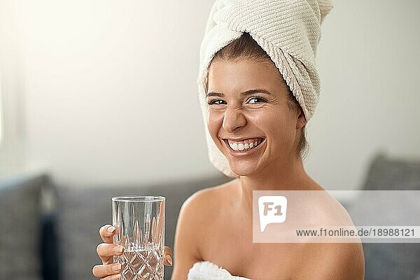 Glückliche attraktive junge Frau mit einem netten freundlichen Grinsen  die ein sauberes weißes Handtuch um ihr Haar trägt und ein Glas reines frisches Wasser hält und in die Kamera lacht