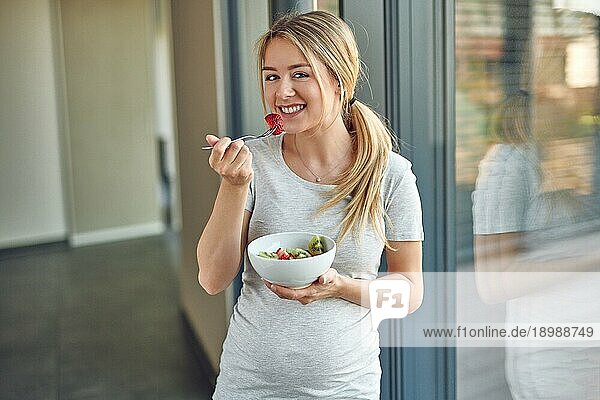 Glückliche  gesunde  schwangere  junge  blonde Frau  die an ein Terrassenfenster gelehnt steht und in die Kamera lächelt  während sie eine Schüssel mit frischem Obstsalat genießt