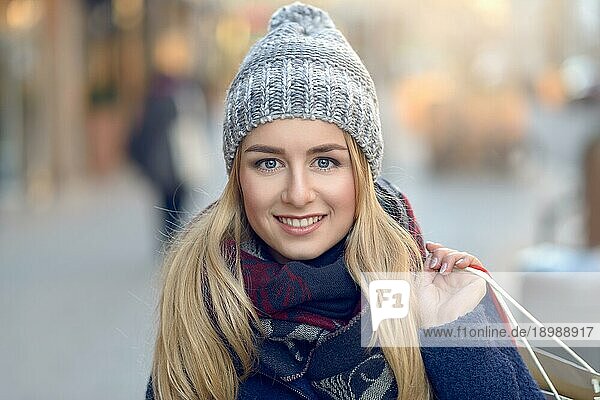 Wunderschöne junge Frau mit Weihnachtseinkäufen in einer gestrickten Wintermütze  die fröhlich lächelt  während sie auf einer städtischen Straße hinter sich blickt