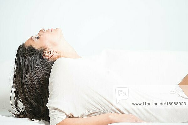 Seitenansicht einer schönen jungen Frau  die sich auf einem Sofa zurücklehnt  den Kopf zurücklehnt und ihr langes brünettes Haar hinter sich herabhängen lässt  während sie sich zu Hause beim Träumen entspannt