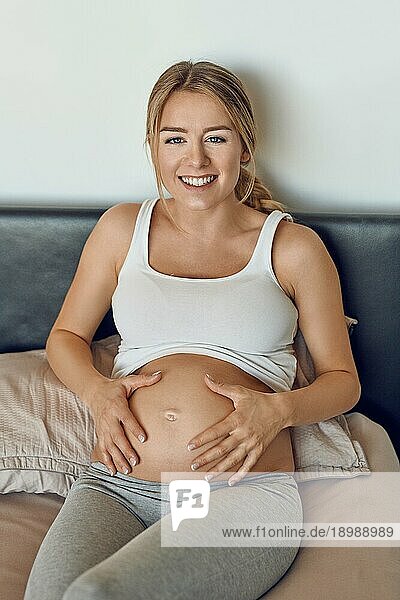 Glücklich lächelnde attraktive junge schwangere Frau  die ihren nackten Babybauch in den Händen wiegt  während sie sich auf einem Bett entspannt und in die Kamera schaut