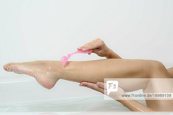 Anmutige junge Frau rasiert die Haut an ihren nackten Beinen in der Badewanne mit einem rosa Einwegrasierer in einem Gesundheits  Schönheits und Körperpflegekonzept