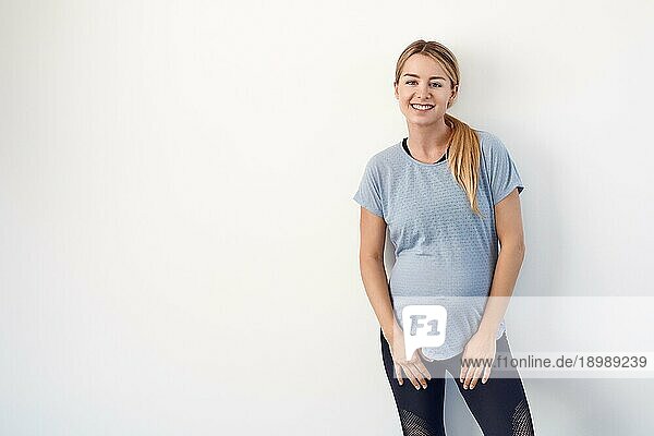 selbstbewusste  entspannte  attraktive  junge  schwangere Frau  die mit verschränkten Armen auf ihrem nackten Babybauch steht und mit einem freundlichen Lächeln in die Kamera blickt  auf weißem Grund mit Leerzeichen