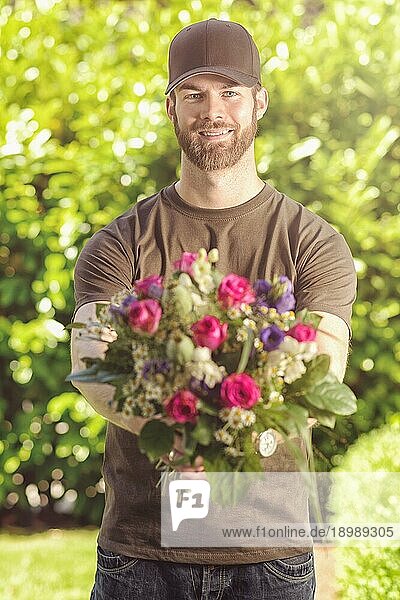 Dreiviertelansicht eines lächelnden  bärtigen Mannes in den 20ern  der eine braune Baseballmütze  ein braunes TShirt und Jeans trägt und einen Blumenstrauß hält