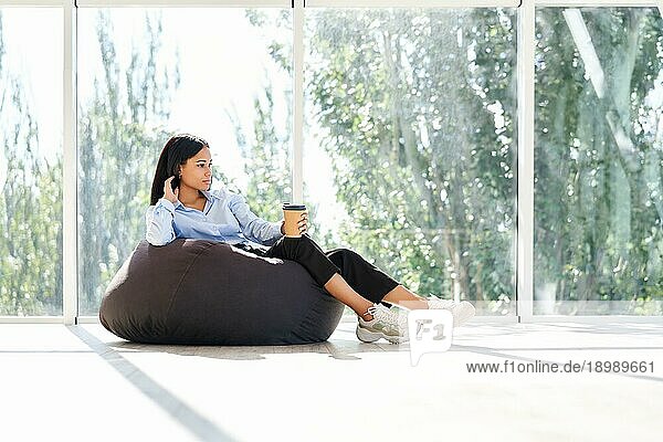 Hübsche afrikanische amerikanische Geschäftsfrau entspannt auf Sitzsack Stuhl mit Tasse Kaffee auf Panoramafenster Hintergrund in modernen kreativen Büro mit Kopie Raum. Pause Zeit Konzept  Geschäftserfolg