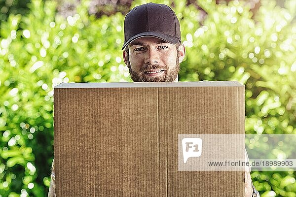 Ein lächelnder Briefträger  der ein großes braunes Papppaket trägt und darauf wartet  eine Lieferung an ein Haus zu machen  während er über die Oberseite des Kartons in die Kamera schaut  vor einem Hintergrund von frischem Frühlingsgrün