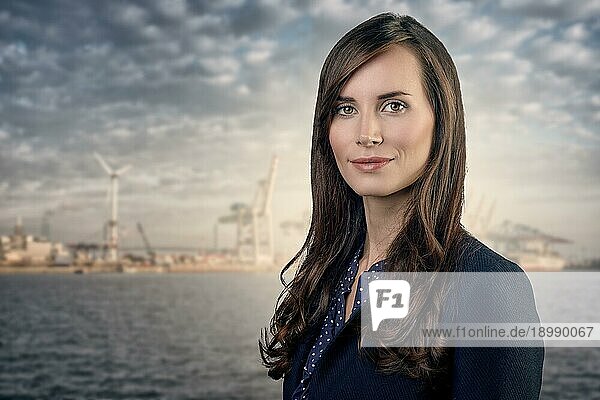 Attraktive Geschäftsfrau mit lockigen braunen Haaren  die eine elegante Jacke trägt  steht vor einem Hafen oder einem Geschäftshafen und lächelt in die Kamera  Oberkörperporträt