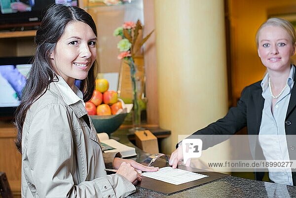 Lächelnder attraktiver junger weiblicher Gast in einer Hotellobby  der sich mit der Rezeptionistin unterhält  als sie bei ihrer Ankunft eincheckt