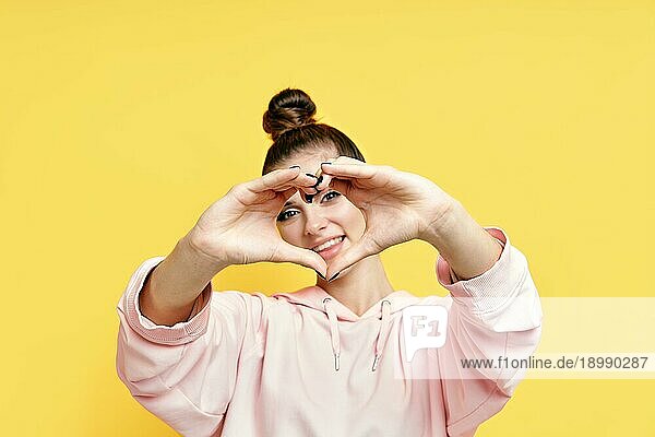Junge lächelnde hübsche Frau macht Herzsymbol mit den Händen auf gelbem Hintergrund. Fokus auf Arns. Körpersprache