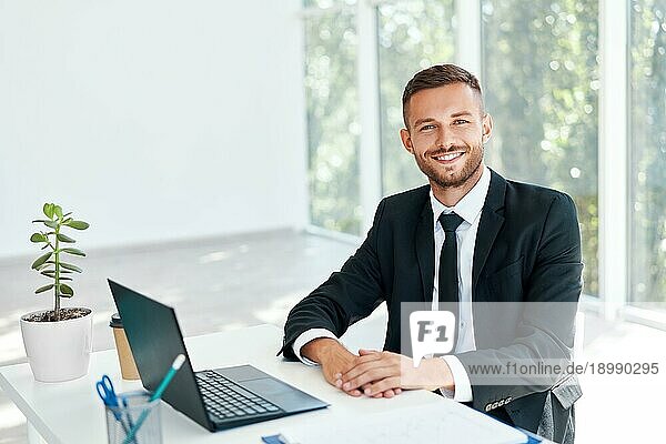 Stilvolle lächelnde Geschäftsmann in eleganten Anzug sitzt an seinem Schreibtisch in einem hellen modernen Büro. geschäftlichen Erfolg Konzept