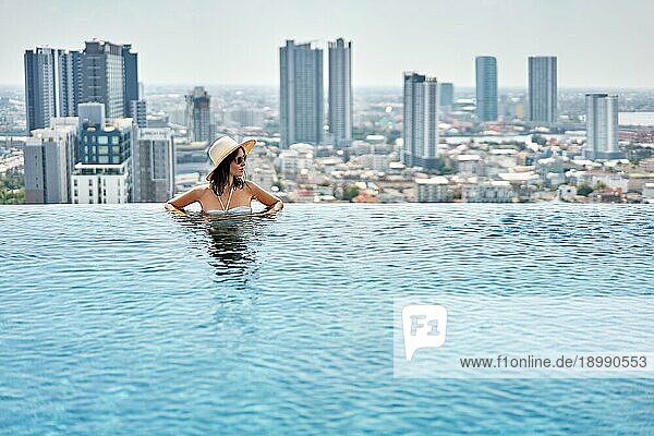 Junge Frau mit Hut genießt Sommerurlaub im Schwimmbad auf dem Dach mit Blick auf die Stadt