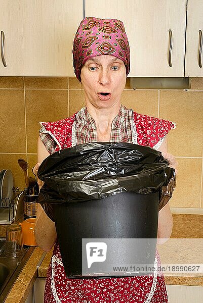 Eine Frau mit Kopftuch und roter Schürze schaut in der Küche in einen schwarzen Mülleimer mit einem Müllsack. Sie ist sehr beunruhigt über den schlechten Geruch
