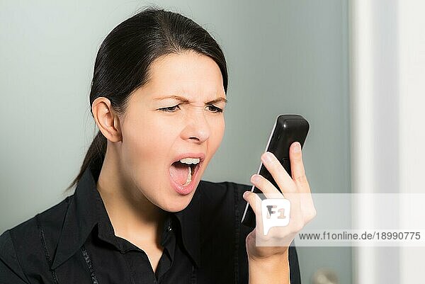 Porträt einer brünetten kaukasischen jungen Frau  die schreit  während sie auf ihr Handy schaut  Symbol für Stress und negative Gefühle  auf grauem Hintergrund
