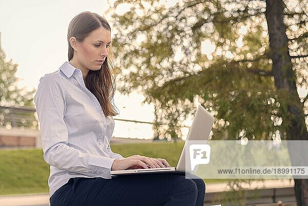 Attraktive junge Frau in formeller Kleidung bei der Arbeit im Freien an einem Laptop  mit einer drahtlosen Verbindung zum Internet  an einem schönen Sommertag