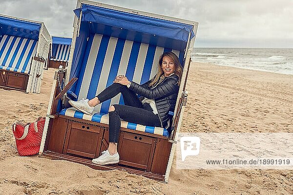 Attraktive schlanke blonde Frau  die in einer Strandhütte mit blauweiß gestreiftem Stoff an einem Sandstrand an einem bewölkten Tag sitzt und in die Kamera lächelt
