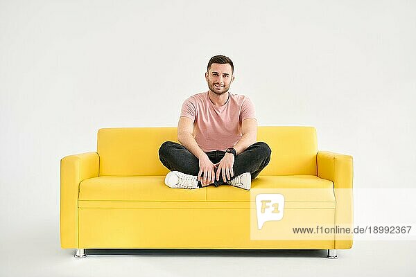 Junger trendiger Mann posiert auf gelbem Sofa sitzend und schaut in die Kamera. Entspannen Konzept