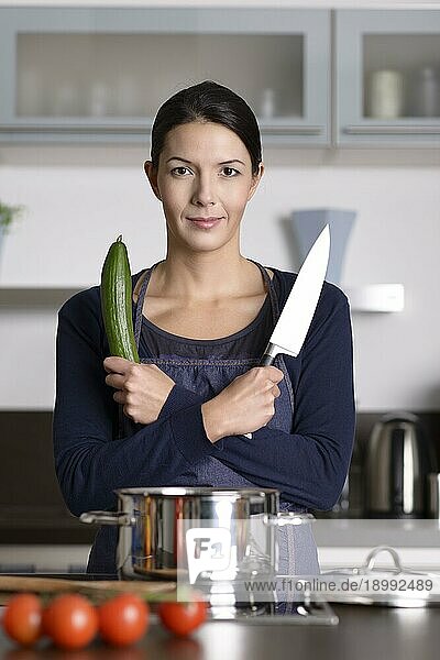 Glückliche junge Frau posiert in ihrer Küche mit verschränkten Armen  ein Messer und frische Gurken haltend  während sie ein gesundes Abendessen zubereitet  Frontalansicht am Herd stehend