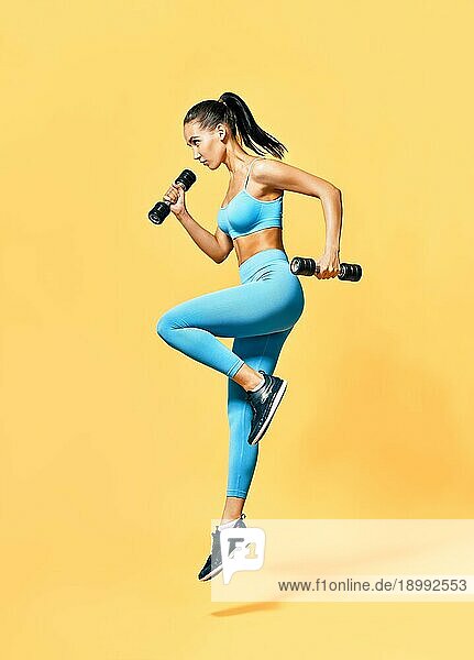 Sportliche Frau in Sportkleidung springt mit Hanteln auf gelbem Hintergrund. Konzept für Stärke und Motivation. Profilporträt in voller Länge