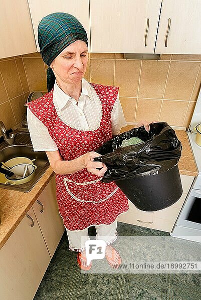 Eine Frau mit Kopftuch und roter Schürze schaut in der Küche in einen schwarzen Mülleimer mit einem Müllsack. Sie ist sehr beunruhigt über den schlechten Geruch