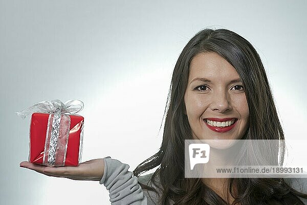 Hübsche junge Frau mit langen brünetten Haaren und einem schönen sanften Lächeln  die ein buntes rotes Geschenk für einen geliebten Menschen überreicht