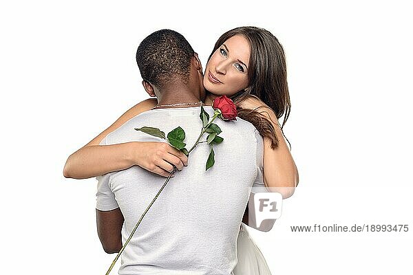 Sentimentale junge Frau  die ihren Freund oder Liebsten umarmt  während sie zärtlich auf eine einzelne rote Rose herablächelt  die er ihr gerade geschenkt hat  symbolisch für Liebe und Romantik oder Valentinstag  gemischtrassiges Paar