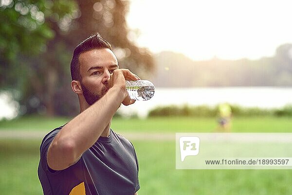 Junger Mann mit Bart  der eine Flasche Wasser trinkt  während er eine von Bäumen gesäumte Allee in einem Park hinuntergeht  Nahaufnahme von Kopf und Schultern