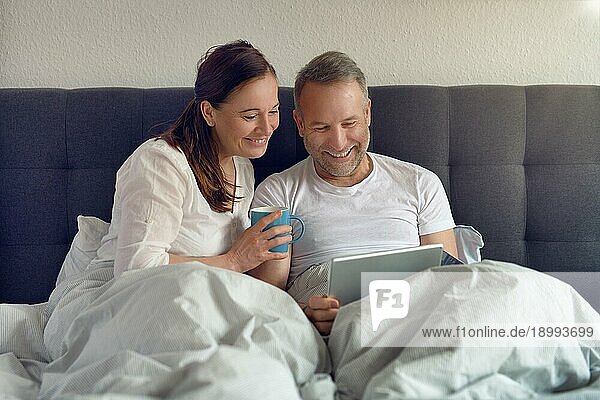 Glückliches Paar mittleren Alters  das morgens im Bett sitzt  Kaffee trinkt und mit einem digitalen Tablet online einkauft