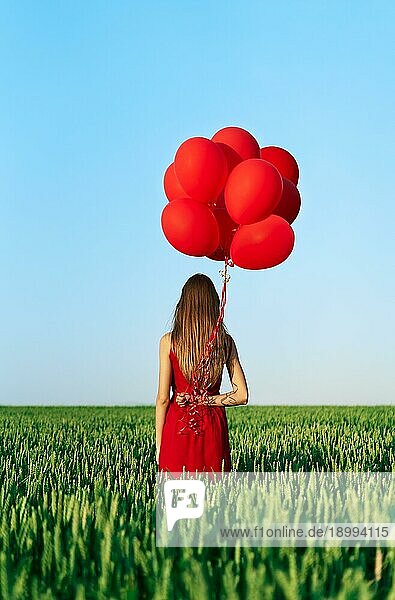 Rückenansicht einer jungen Frau in rotem Kleid  die mit roten Luftballons auf einer grünen Sommerwiese steht. Spaß  Glück  Feier Konzept