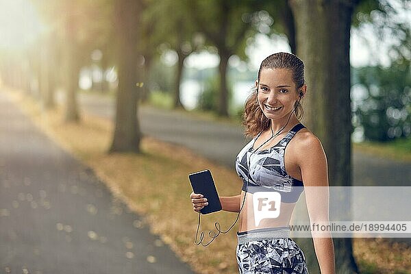 Attraktive sportliche Frau  die mit ihrem Handy Musik hört  joggt auf einer von Bäumen gesäumten Straße und lächelt fröhlich in die Kamera