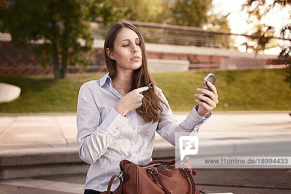 Junge Frau beim Auffrischen ihrer Frisur in einer städtischen Straße  die sich selbst in einem kleinen Handspiegel betrachtet  während sie ihr langes braunes Haar kämmt